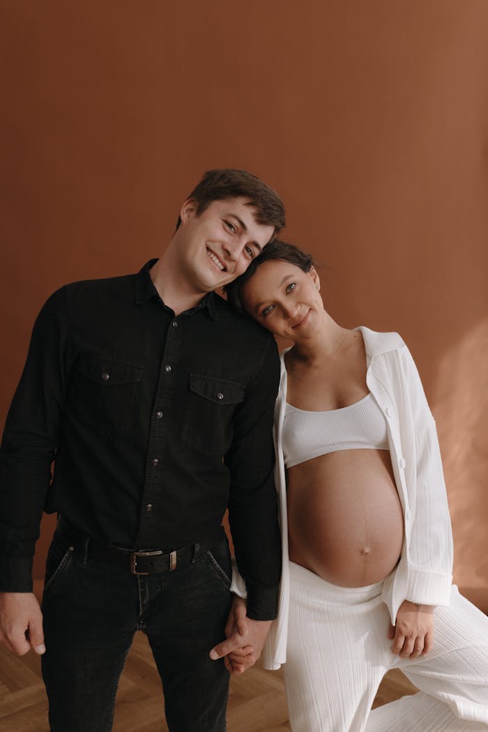 A joyful expecting couple smiling together in a studio with a brown background.